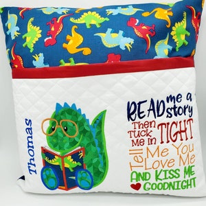 May include: A white pillow with a pocket for books, featuring a green dinosaur wearing glasses and reading a book. The pillow has a red and blue dinosaur print on the back and the text "READ me a story Then TUCK me in Tell Me You LOVE Me AND KISS ME GOODNIGHT" embroidered on the front.