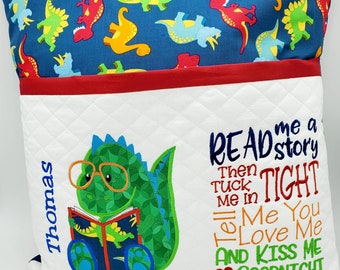 Pode incluir: Uma almofada branca com um bolso para livros, apresentando um dinossauro verde usando óculos e lendo um livro. A almofada tem uma estampa de dinossauros vermelho e azul nas costas e o texto "READ me a story Then TUCK me in Tell Me You LOVE Me AND KISS ME GOODNIGHT" bordado na frente.