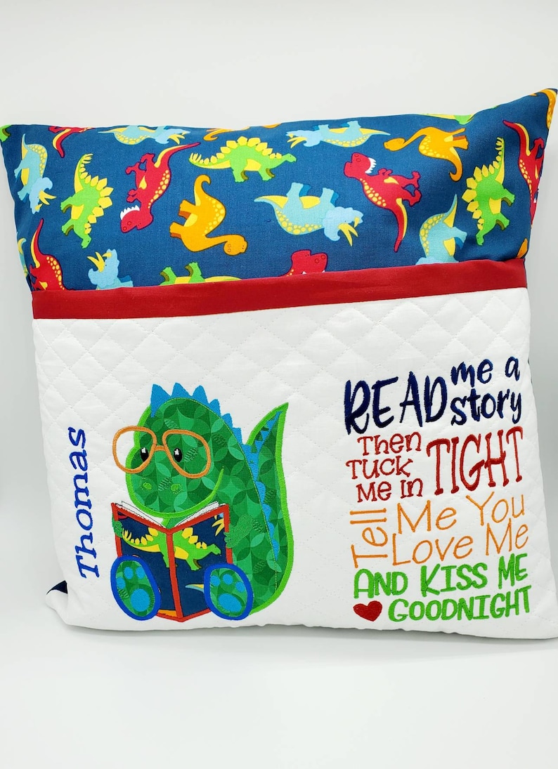 May include: A white pillow with a pocket for books, featuring a green dinosaur wearing glasses and reading a book. The pillow has a red and blue dinosaur print on the back and the text "READ me a story Then TUCK me in Tell Me You LOVE Me AND KISS ME GOODNIGHT" embroidered on the front.