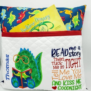 May include: A white pillow with a pocket on the front. The pocket has a red border and a yellow book inside. The pillow has a green dinosaur with glasses reading a book on the left side. The right side of the pillow has the text "READ me a story Then TUCK me in TIGHT Tell Me You LOVE Me AND KISS ME GOODNIGHT". The pillow is personalized with the name "Thomas" on the left side.