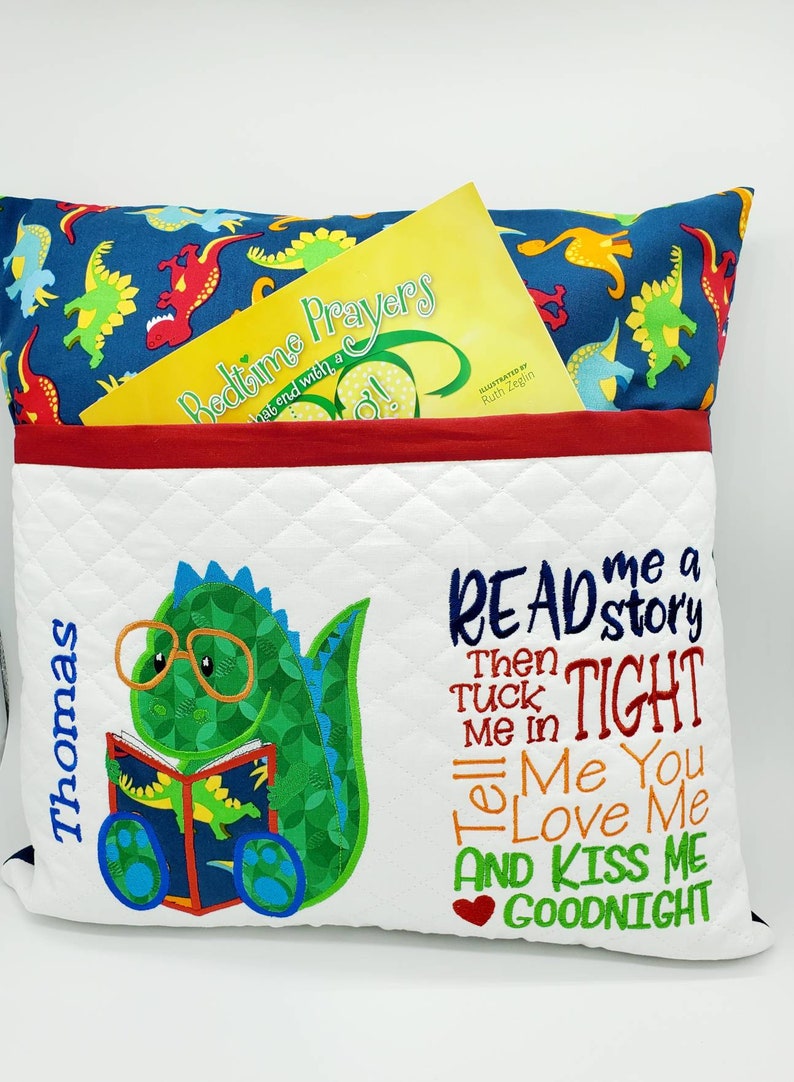 May include: A white pillow with a pocket on the front. The pocket has a red border and a yellow book inside. The pillow has a green dinosaur with glasses reading a book on the left side. The right side of the pillow has the text "READ me a story Then TUCK me in TIGHT Tell Me You LOVE Me AND KISS ME GOODNIGHT". The pillow is personalized with the name "Thomas" on the left side.