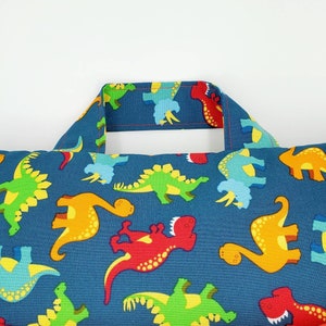 May include: A blue pillow with a dinosaur pattern. The pillow has a handle on the top and is filled with stuffing. The dinosaurs are in various colors, including red, green, yellow, and orange.