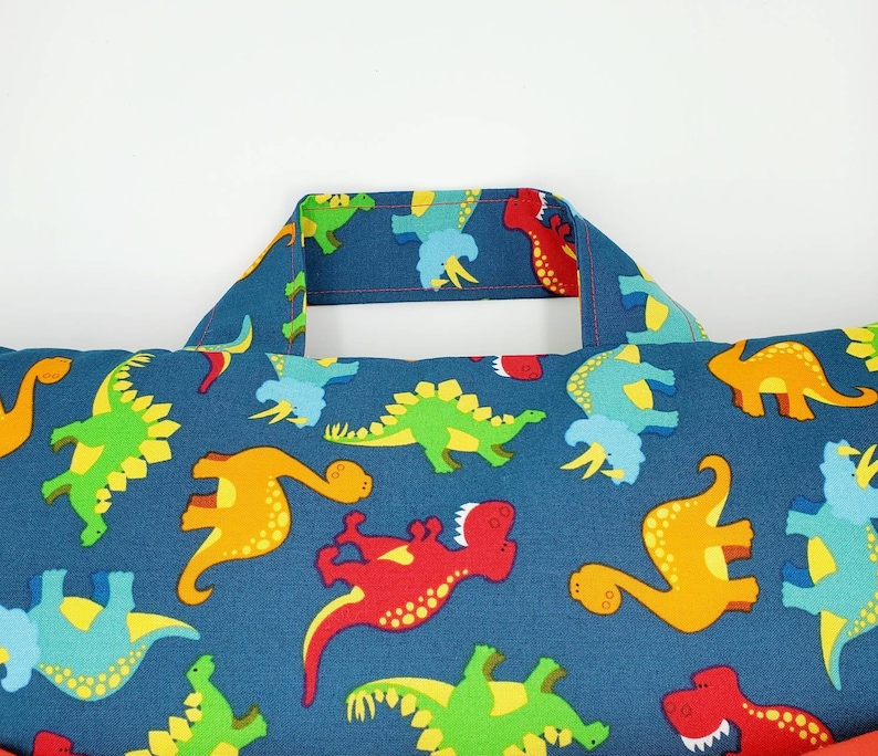 May include: A blue pillow with a dinosaur pattern. The pillow has a handle on the top and is filled with stuffing. The dinosaurs are in various colors, including red, green, yellow, and orange.