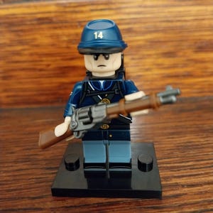 May include: A blue and black Lego minifigure of a soldier wearing a blue hat with the number "14" on it. The figure is holding a brown rifle and is standing on a black base.
