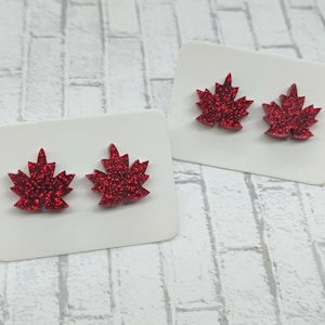 May include: A pair of red glitter maple leaf stud earrings. The earrings are on a white background.