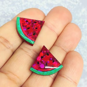 May include: Two watermelon slice-shaped earrings. The earrings have a red glitter interior with black and pink accents, a green rind, and a white border. The earrings are held in a person's hand.