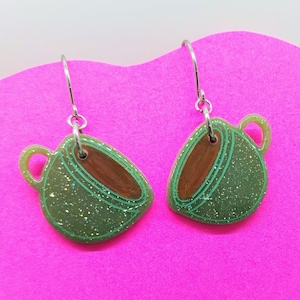 May include: A pair of green glittery coffee cup earrings with silver hooks. The earrings have a brown interior and a green outline. The background is a bright pink.