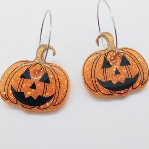 Puede incluir: Un par de pendientes de calabaza naranja con purpurina y caras de calabaza negras. Los pendientes son de aro con aros plateados.