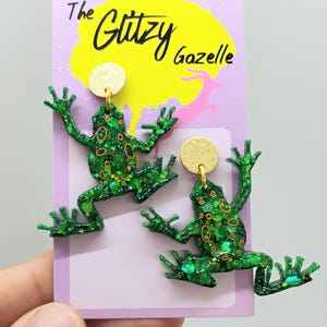 May include: Green frog-shaped earrings with glitter and orange detailing, attached to gold-coloured circular studs. The earrings are displayed on a purple card with the text "The Glitzy Gazelle".
