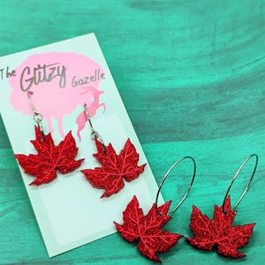 May include: Red glitter maple leaf earrings with silver hooks and a pair of red glitter maple leaf hoop earrings. The earrings are displayed on a white card with the text "The Glitzy Gazelle".