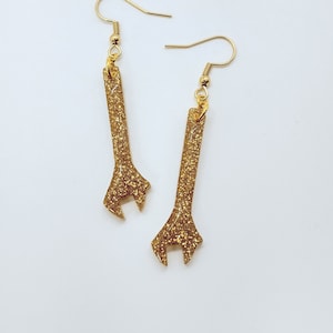 May include: A pair of gold glitter wrench earrings. The earrings are made of metal and have a gold glitter finish.
