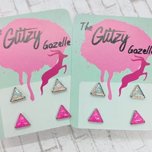 Puede incluir: Dos pares de pendientes de botón en tarjetas de exhibición. Cada tarjeta presenta un diseño de salpicaduras rosas con el texto "The Glitzy Gazelle" y una silueta de gacela rosa. Los pendientes son triangulares, con opciones de purpurina plateada y rosa.