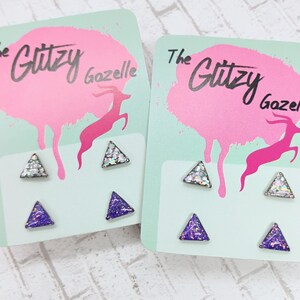 May include: Two sets of triangle stud earrings on cards. Each card has a pink splattered design with a pink gazelle and the text "The Glitzy Gazelle". One set has silver glitter triangles, the other has purple glitter triangles.