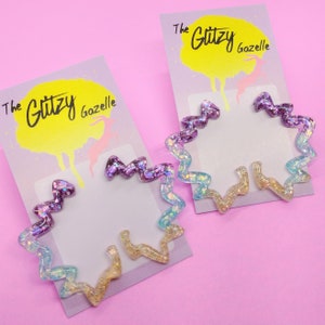 May include: Two pairs of iridescent glitter hoop earrings. One pair is purple and blue, the other is gold and blue. The earrings are displayed on a purple and yellow background with the text "The Glitzy Gazelle".