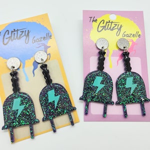 May include: A pair of dangle earrings featuring a black plug with a green lightning bolt design. The earrings are made with glitter and have a silver accent at the top. The earrings are displayed on a pink and yellow card with the text "The Glitzy Gazelle".