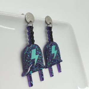 Puede incluir: Un par de pendientes de purpurina morada con forma de enchufe el&eacute;ctrico con un dise&ntilde;o de rayo turquesa.