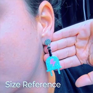 Puede incluir: Un par de pendientes de color azul turquesa con un dise&ntilde;o de rayo rosa. Los pendientes cuelgan de una cadena negra y tienen un cierre plateado.