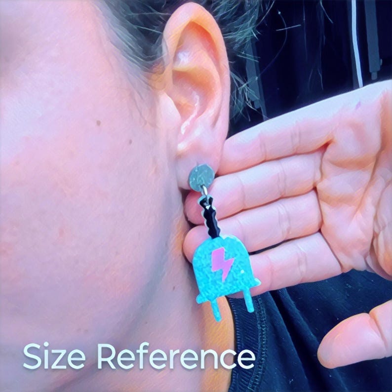 Puede incluir: Un par de pendientes de color azul turquesa con un dise&ntilde;o de rayo rosa. Los pendientes cuelgan de una cadena negra y tienen un cierre plateado.