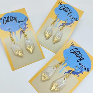 May include: A pair of white and gold resin earrings with a geometric shape. The earrings are on a white card with the text "The Glitzy Gazelle" in blue and yellow.