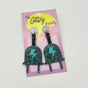 Puede incluir: Un par de pendientes de purpurina negra y verde con un dise&ntilde;o de rayo turquesa. Los pendientes est&aacute;n en una tarjeta rosa con el texto "The Glitzy Gazelle".