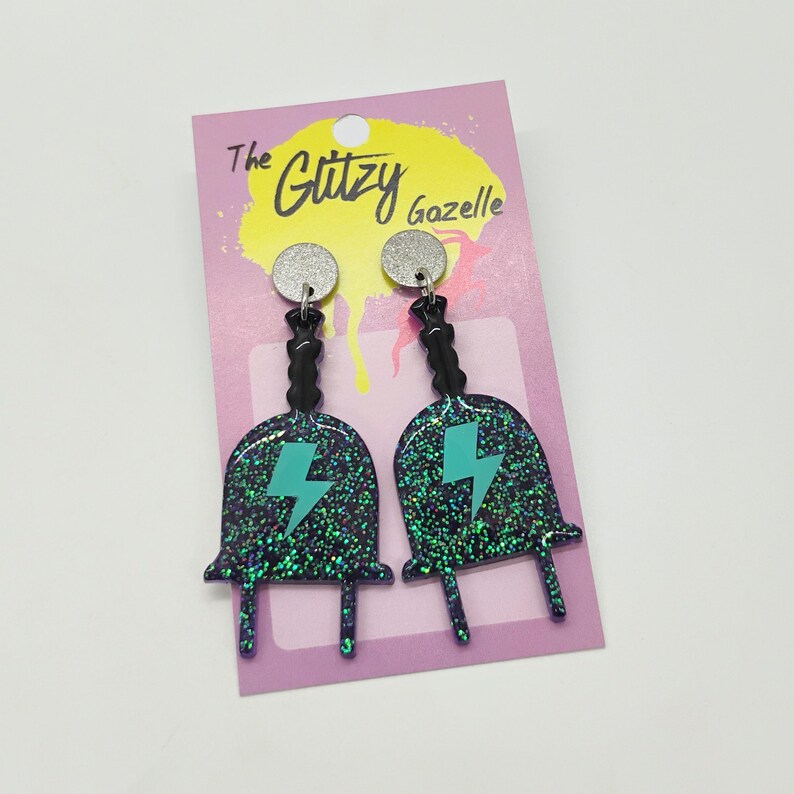 Puede incluir: Un par de pendientes de purpurina negra y verde con un dise&ntilde;o de rayo turquesa. Los pendientes est&aacute;n en una tarjeta rosa con el texto "The Glitzy Gazelle".
