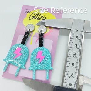 Puede incluir: Un par de pendientes de color azul turquesa con un dise&ntilde;o de rayo rosa y detalles negros. Los pendientes miden aproximadamente 5 cm de largo y se muestran junto a una regla para referencia de tama&ntilde;o. Los pendientes est&aacute;n hechos con purpurina y tienen un acabado brillante.