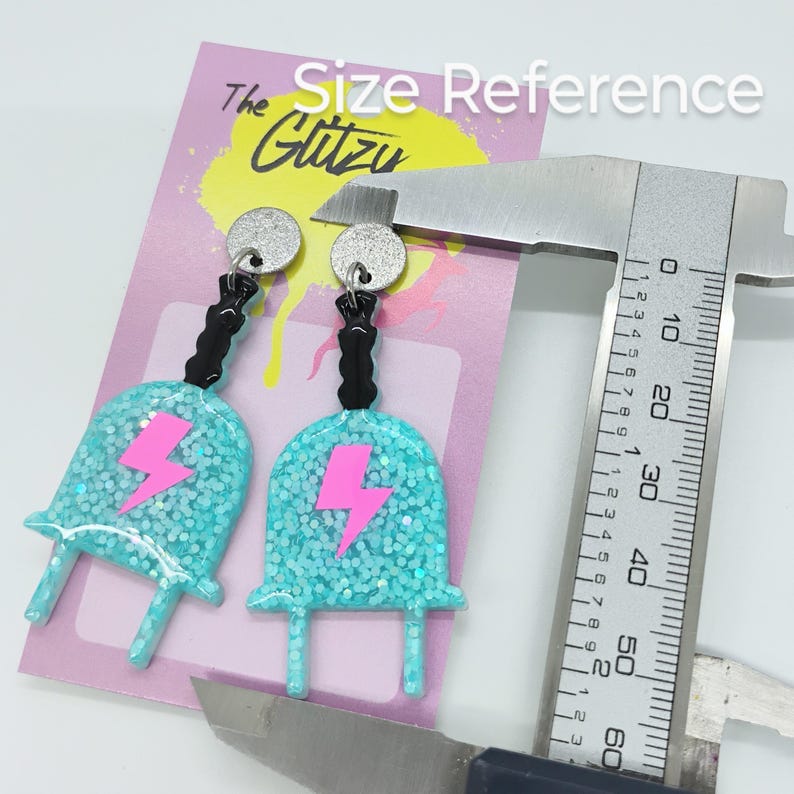 Puede incluir: Un par de pendientes de color azul turquesa con un dise&ntilde;o de rayo rosa y detalles negros. Los pendientes miden aproximadamente 5 cm de largo y se muestran junto a una regla para referencia de tama&ntilde;o. Los pendientes est&aacute;n hechos con purpurina y tienen un acabado brillante.
