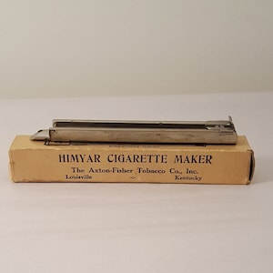 May include: A brown cardboard box with the text "HIMYAR CIGARETTE MAKER" and "The Axion-Fisher Tobacco Co., Inc. Louisville, Ky. Kentucky" printed on it. The box contains a silver cigarette maker.