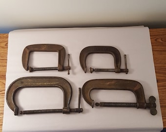 May include: Four metal C-clamps, two with the word "Adjustable" printed on them, on a white surface.