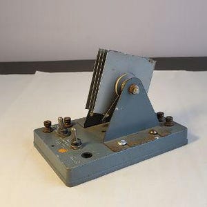May include: A vintage gray metal device with a series of switches, knobs, and a hinged panel. The panel has a stack of metal plates. The device is set on a white surface.