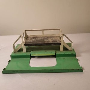 May include: A white metal toy playset with a green platform and a raised platform with a white railing. The playset is made of metal and has a simple design.