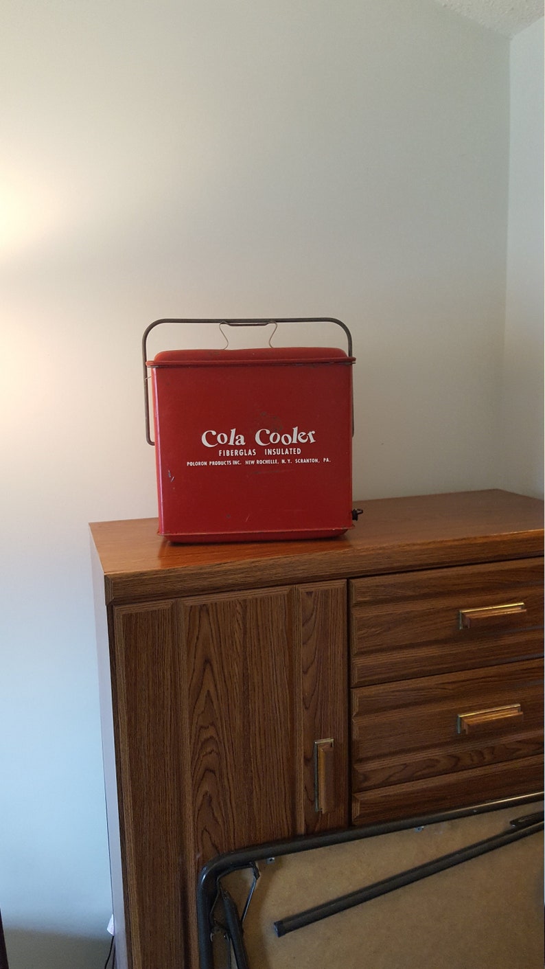 May include: Red vintage cooler with a white "Cola Coolers Fiberglass Insulated" label. The cooler has a metal handle and is sitting on a wooden cabinet.