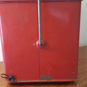 May include: Red metal cooler with a carrying handle and a latch on the front. The cooler has a worn, vintage look.
