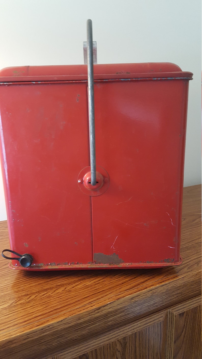 May include: Red metal cooler with a carrying handle and a latch on the front. The cooler has a worn, vintage look.