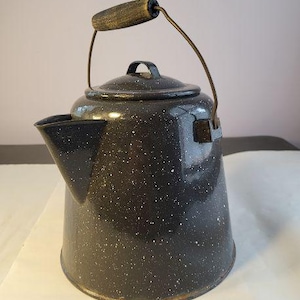 May include: A speckled black enamelware kettle with a spout, lid, and handle. The handle is made of wood and metal. The kettle is a classic design, likely for camping or outdoor use. The kettle is approximately 10 inches tall.