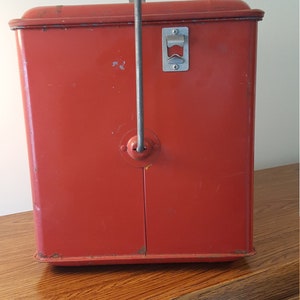 May include: Red metal cooler with a built-in bottle opener and a handle. The cooler is sitting on a wooden surface.