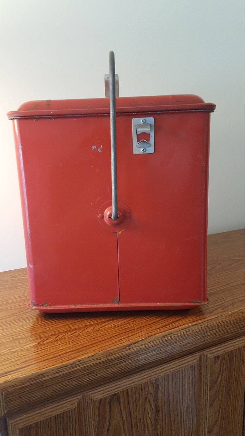 May include: Red metal cooler with a built-in bottle opener and a handle. The cooler is sitting on a wooden surface.
