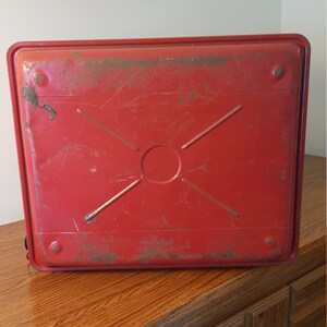 May include: Red metal tray with a cross pattern and a circular indentation in the center. The tray has a worn, vintage look.