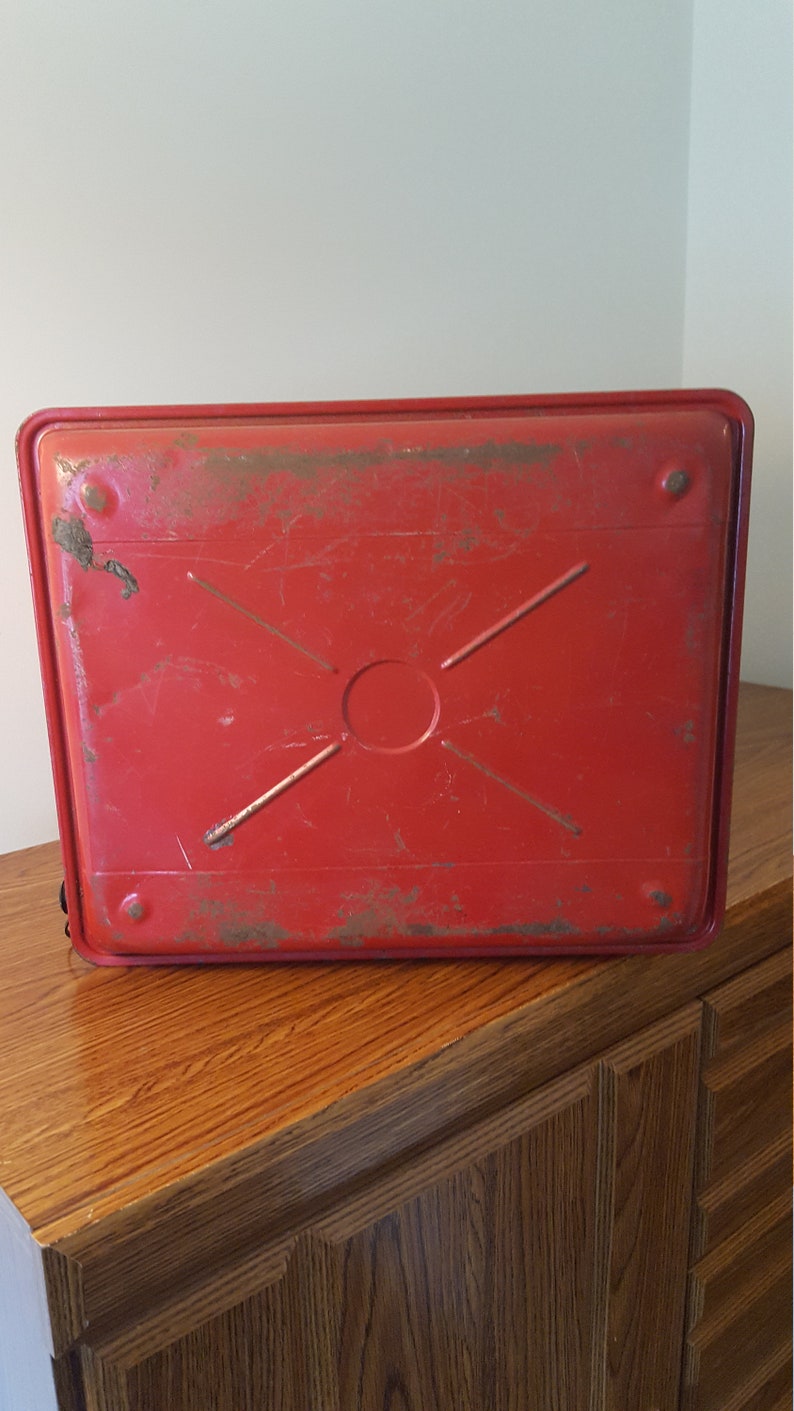 May include: Red metal tray with a cross pattern and a circular indentation in the center. The tray has a worn, vintage look.