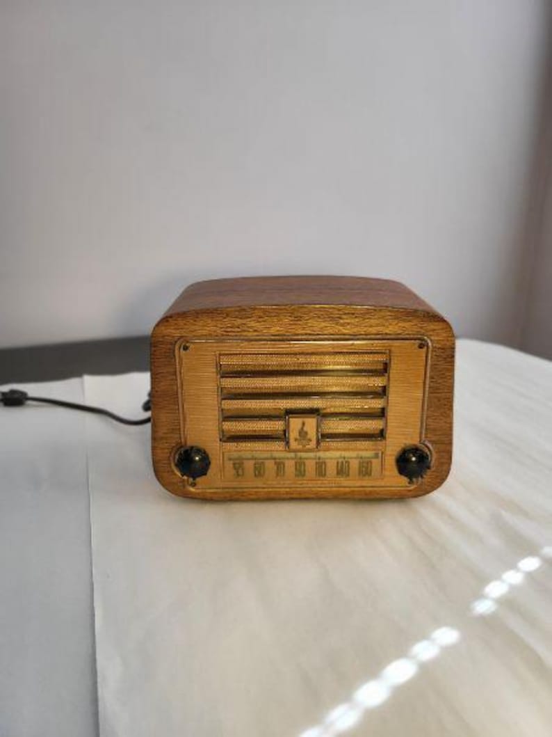 K&ouml;nnte beinhalten: Ein Vintage-Holzradio mit goldfarbenem Lautsprechergrill und Abstimmrad. Das Radio hat eine dunkle Holzoberfl&auml;che und zwei schwarze Kn&ouml;pfe. Das Zifferblatt zeigt Zahlen von 55 bis 160 an. Ein schwarzes Netzkabel erstreckt sich von der R&uuml;ckseite.