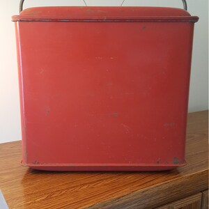May include: A red metal cooler with a metal handle. The cooler is sitting on a wooden surface.