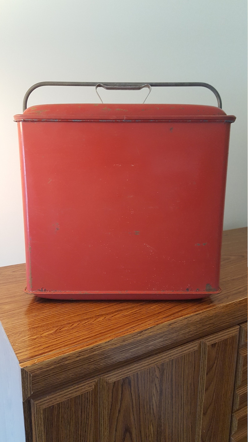 May include: A red metal cooler with a metal handle. The cooler is sitting on a wooden surface.