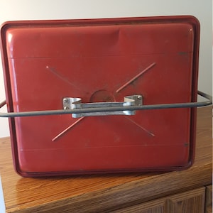 May include: Red metal container with a carrying handle and a metal frame. The container has a slightly textured surface and is sitting on a wooden surface.