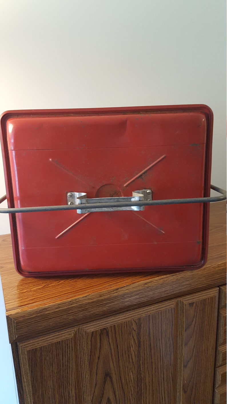 May include: Red metal container with a carrying handle and a metal frame. The container has a slightly textured surface and is sitting on a wooden surface.