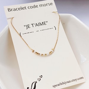 Può includere: Una delicata collana d'oro con un messaggio in codice Morse che dice "JE T'AIME" (Ti amo). La collana presenta piccole perline d'oro e bianche. La carta recita "Bracelet code morse" e include una piccola illustrazione di un uccello.