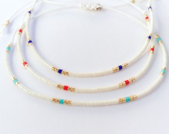 May include: Three white beaded bracelets with gold, red, blue, and turquoise accents. The bracelets are layered on top of each other.