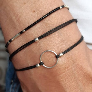 Puede incluir: Tres pulseras en una muñeca. La pulsera superior es una pulsera de cuentas negras y plateadas. Las dos pulseras del medio son cordones negros con cuentas plateadas y un dije circular plateado.