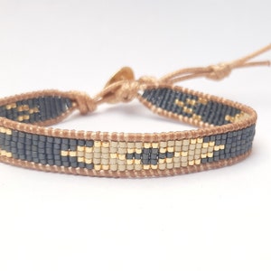 May include: A beaded bracelet with a geometric pattern in black, gold, and beige colors. The bracelet is made with small beads and has a brown cord closure.