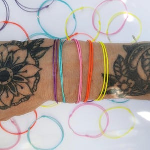 Può includere: Una collezione di braccialetti di filo colorati in viola, turchese, rosa, arancione e giallo. Diversi braccialetti sono indossati su un polso con un tatuaggio floreale. Ulteriori braccialetti in vari colori sono sparsi su una superficie bianca.