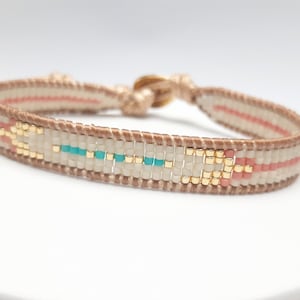 Peut inclure: Un bracelet en perles beige avec un motif de flèche rose et turquoise. Le bracelet a un fermoir doré.
